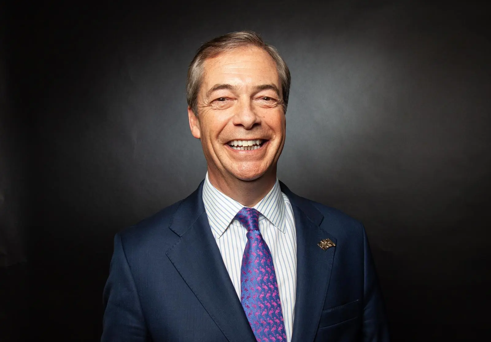 Photo of a smiling man in a blue suit with a colorful tie against a dark background.